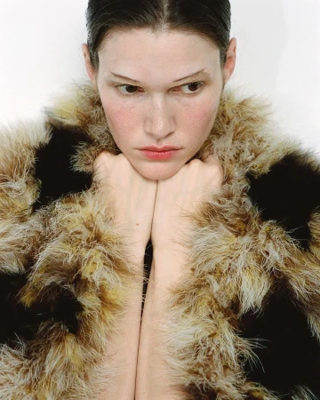 Portrait of a fair-skinned young woman with her chin resting on her hands and wearing a fuzzy, brown and beige faux fur coat in a studio.
