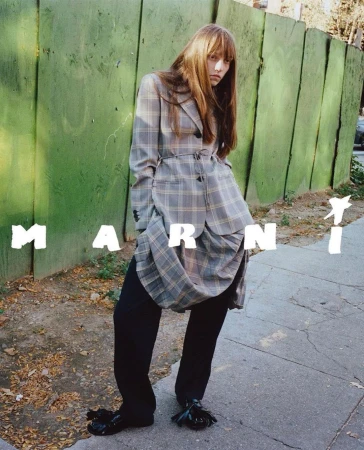 A woman poses in an outdoor urban environment, wearing a plaid jacket and black pants, with the MARNI logo visible in the foreground.
