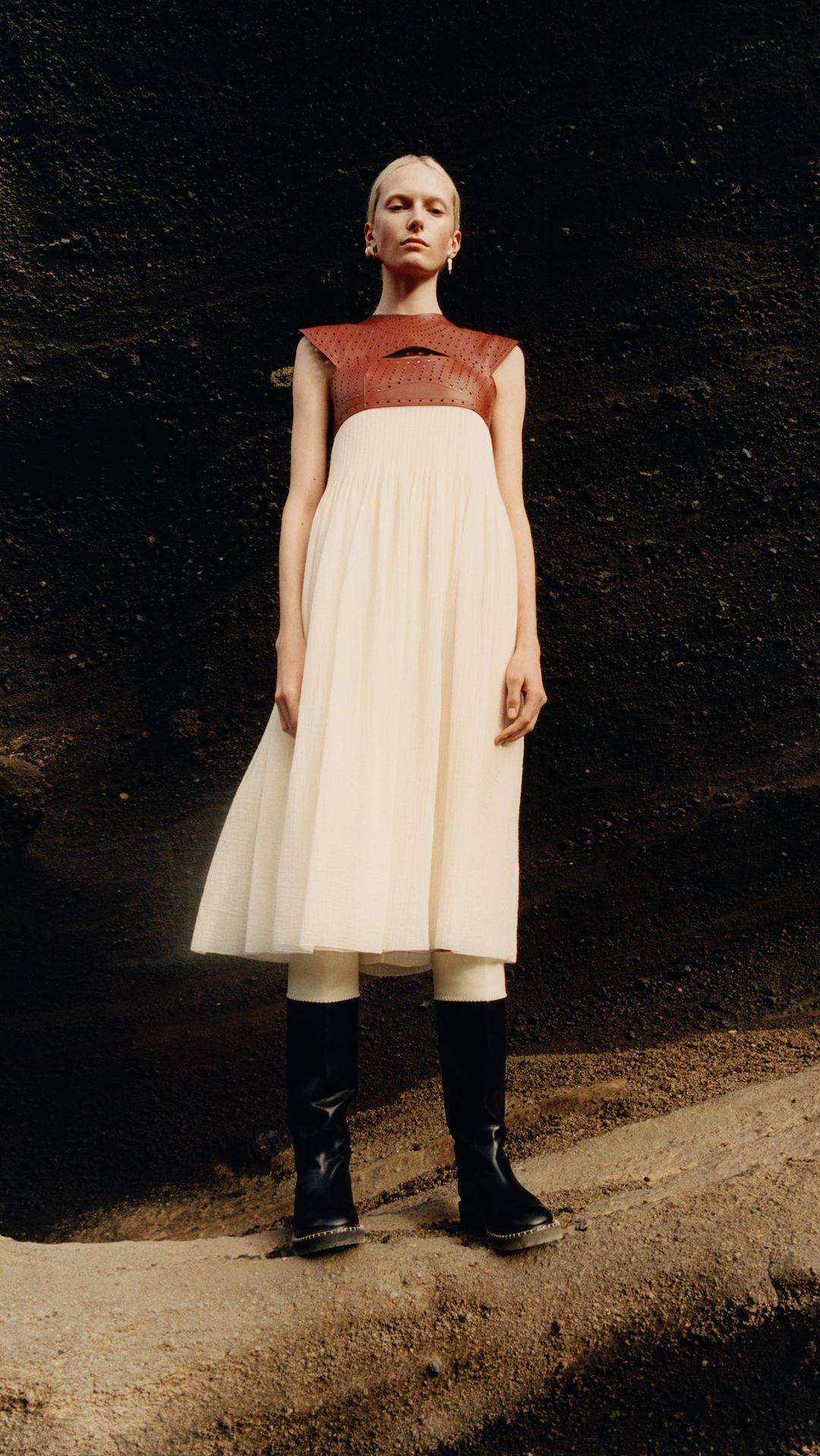 A woman in a light-colored dress and black boots stands in front of a dark wall, photographed in a natural outdoor setting. Fashion photography.