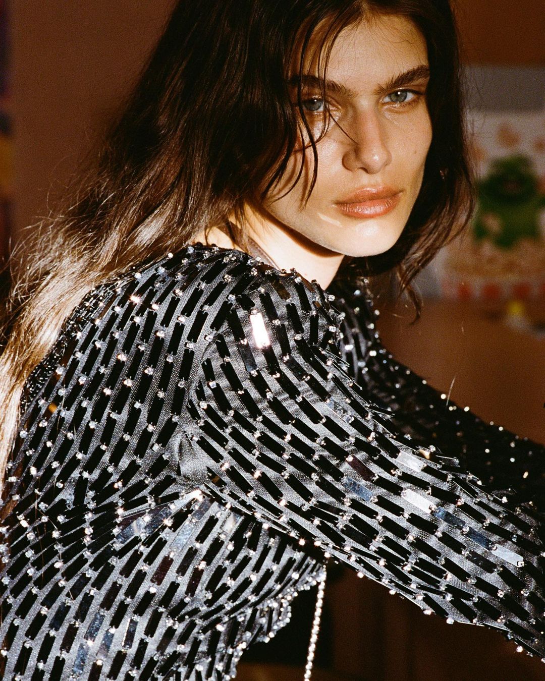 Close-up portrait of a woman with long brown hair wearing a long-sleeved, black sequined dress in an indoor environment with diffused lighting.