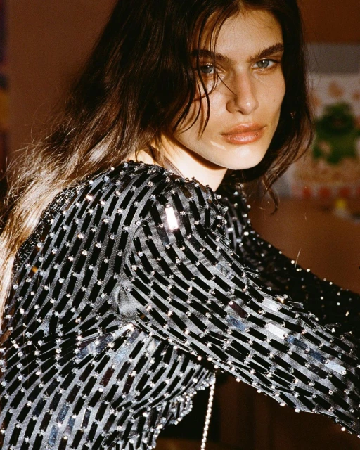 Close-up portrait of a woman with long brown hair wearing a long-sleeved, black sequined dress in an indoor environment with diffused lighting.