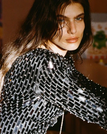 Close-up portrait of a woman with long brown hair wearing a long-sleeved, black sequined dress in an indoor environment with diffused lighting.