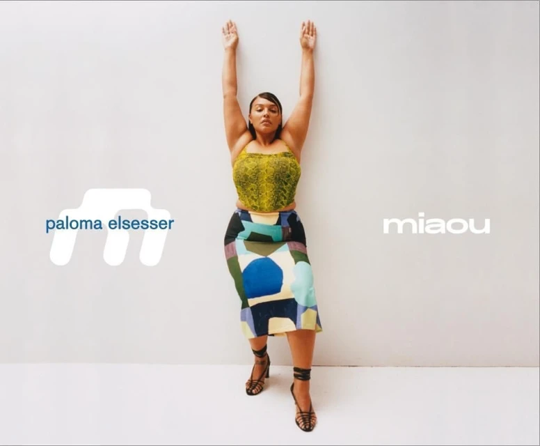 Indoor studio fashion shot of an adult female in a patterned top and skirt with hands raised against a white wall with brand logos.