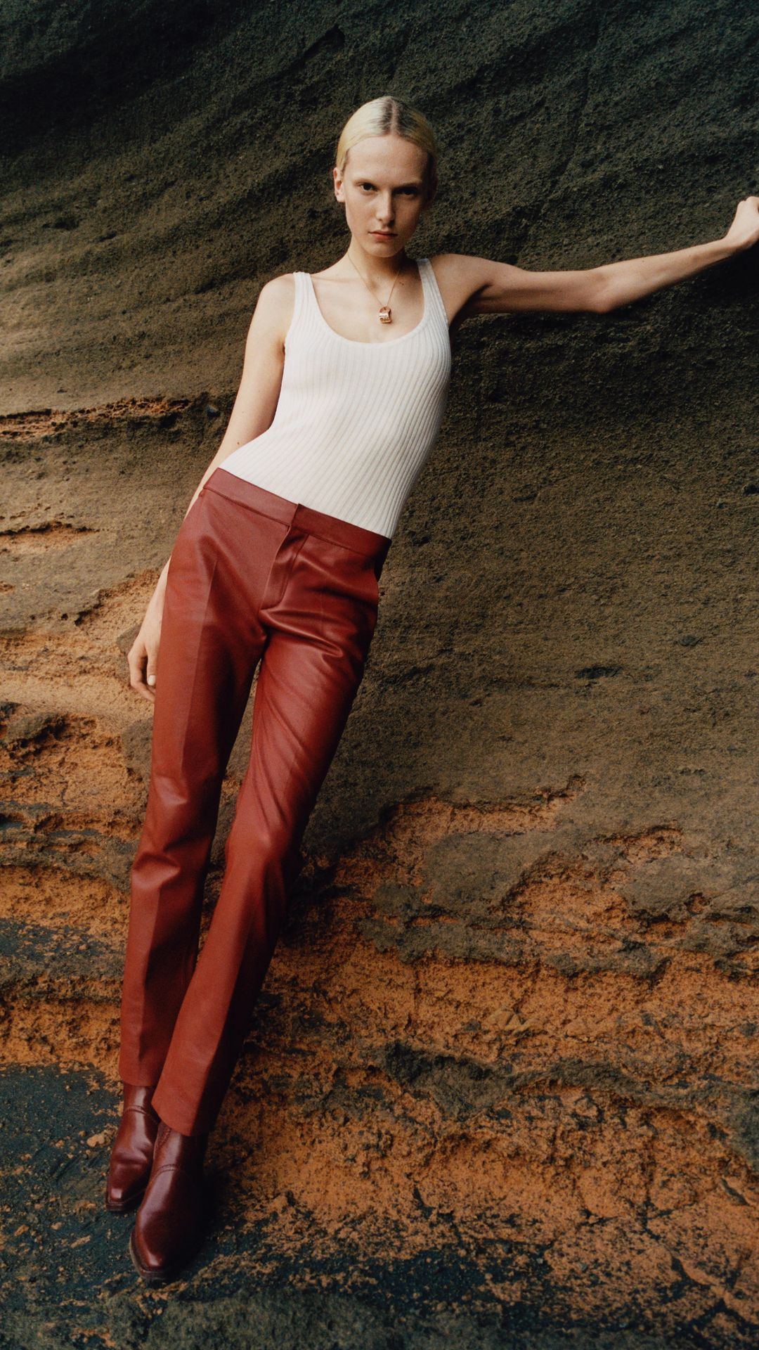 Woman in white top and maroon leather pants poses against a rocky background, captured in natural lighting for a fashion editorial.