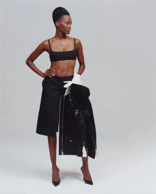 An African American woman poses in a black top, skirt, and heels, wearing white gloves and holding a black jacket in a photo studio.