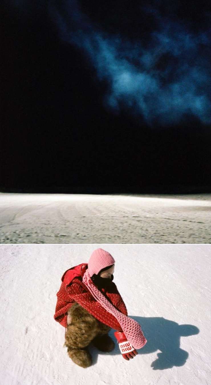 Child in a red jacket sits on snow, hugging a teddy bear against a dark night sky and snow-covered ground. Knitted pink scarf and hat visible.