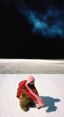 Child in a red jacket sits on snow, hugging a teddy bear against a dark night sky and snow-covered ground. Knitted pink scarf and hat visible.