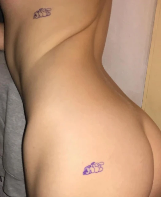 An adult female is lying down with two tattoos, one on her upper back and one on her lower back. She is indoors with fair skin.