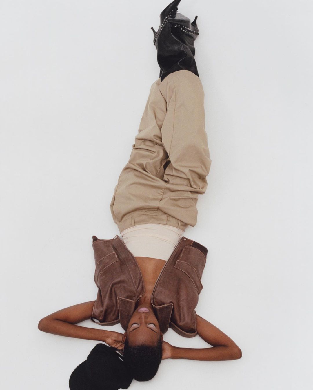 A person wearing a brown vest and khaki pants lies on a white background with their legs raised and pointing upwards.