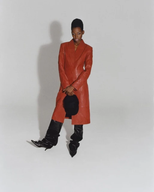 An adult female poses in a red leather coat and black shiny boots against a neutral background, holding a round, soft hat.