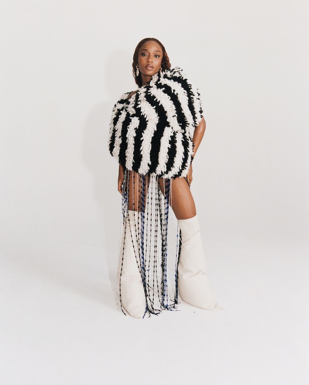 Woman in oversized striped fringe dress and knee-high boots poses in a photo studio against a solid white backdrop, looking at the camera.