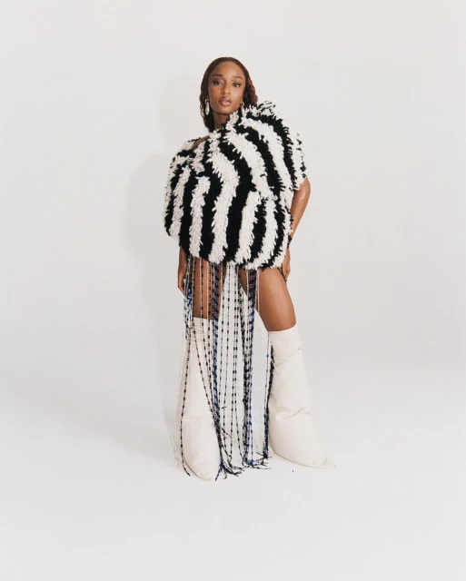 Woman in oversized striped fringe dress and knee-high boots poses in a photo studio against a solid white backdrop, looking at the camera.