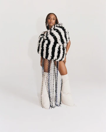 Woman in oversized striped fringe dress and knee-high boots poses in a photo studio against a solid white backdrop, looking at the camera.