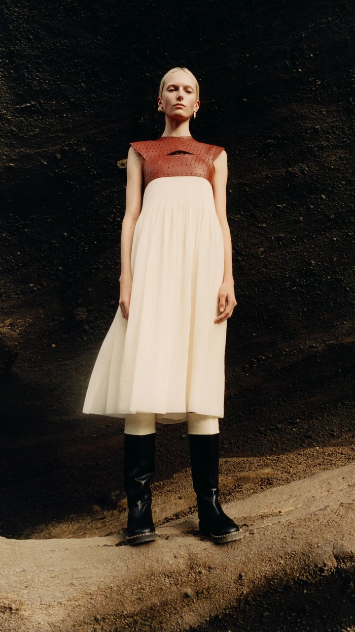 Model standing outdoors wearing a white pleated dress with brown leather bodice and black boots against dark rock.