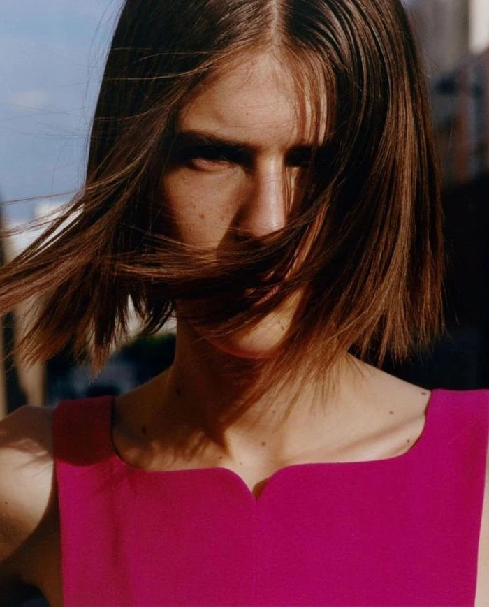 Close-up portrait of a woman with short brown hair blowing across her face wearing a magenta top, shot by Zora Sicher.