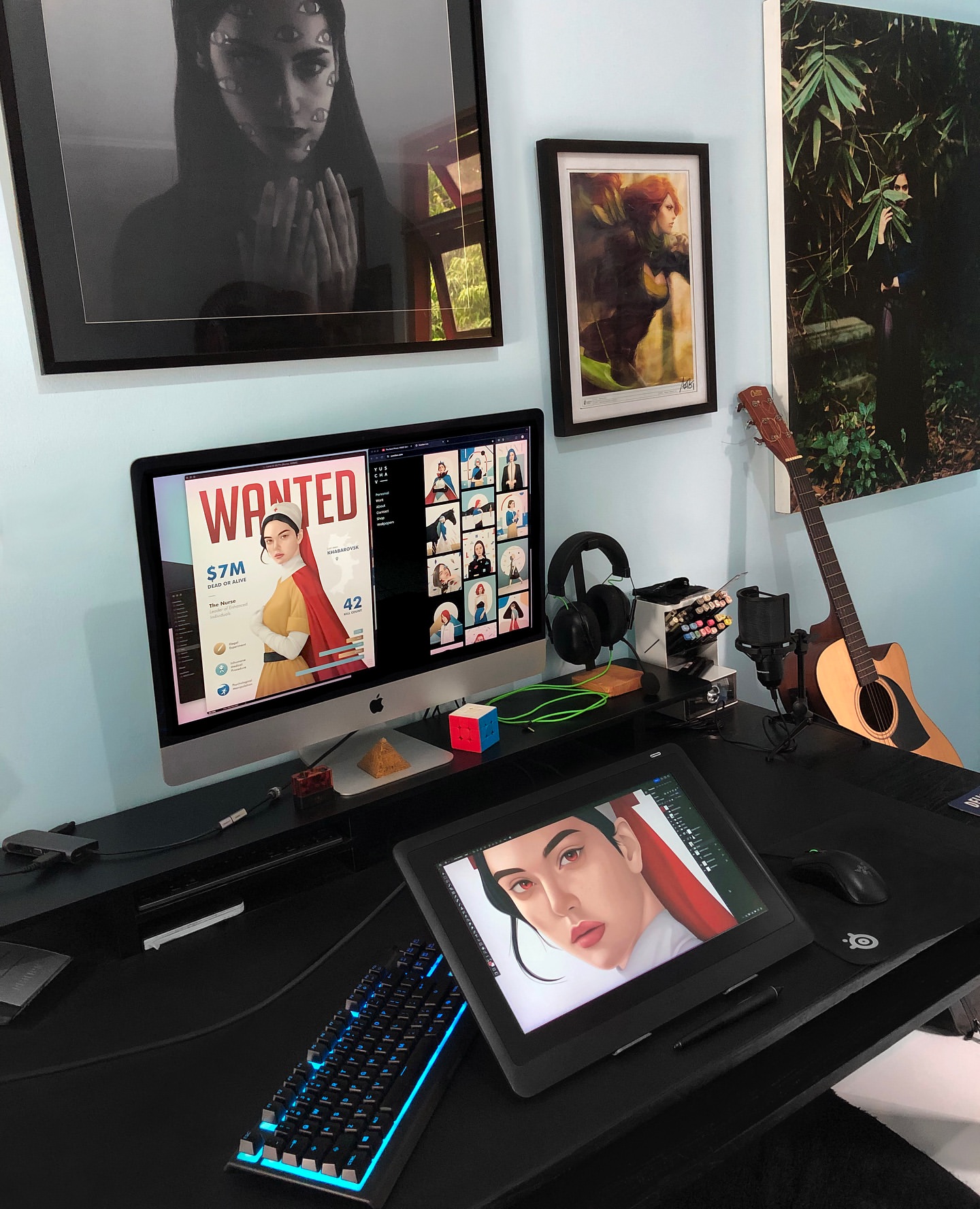 Workspace setup with computer screens displaying creative content, surrounded by art, keyboard, drawing tablet, and artistic tools.