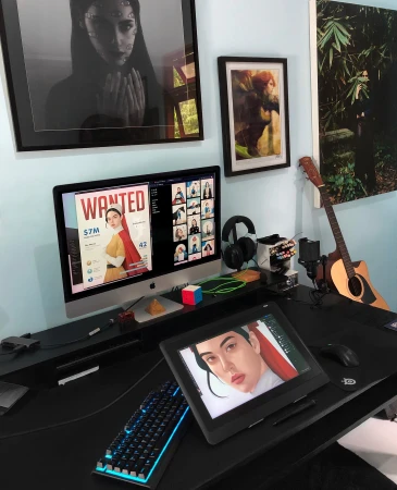 Workspace setup with computer screens displaying creative content, surrounded by art, keyboard, drawing tablet, and artistic tools.