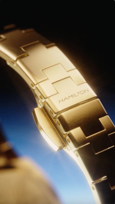 Close-up of a gold-tone metal Hamilton watch band. The band features a clasp with engraved branding, set against a dark blue background.