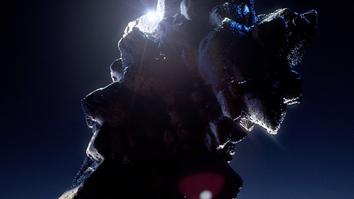 Abstract dark rock formation with textured surfaces and bright lighting, illuminated against a dark blue gradient background.