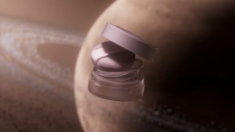 Eye shadow compact in a clear jar with a lavender lid is showcased against a blurry backdrop of a planet with rings.