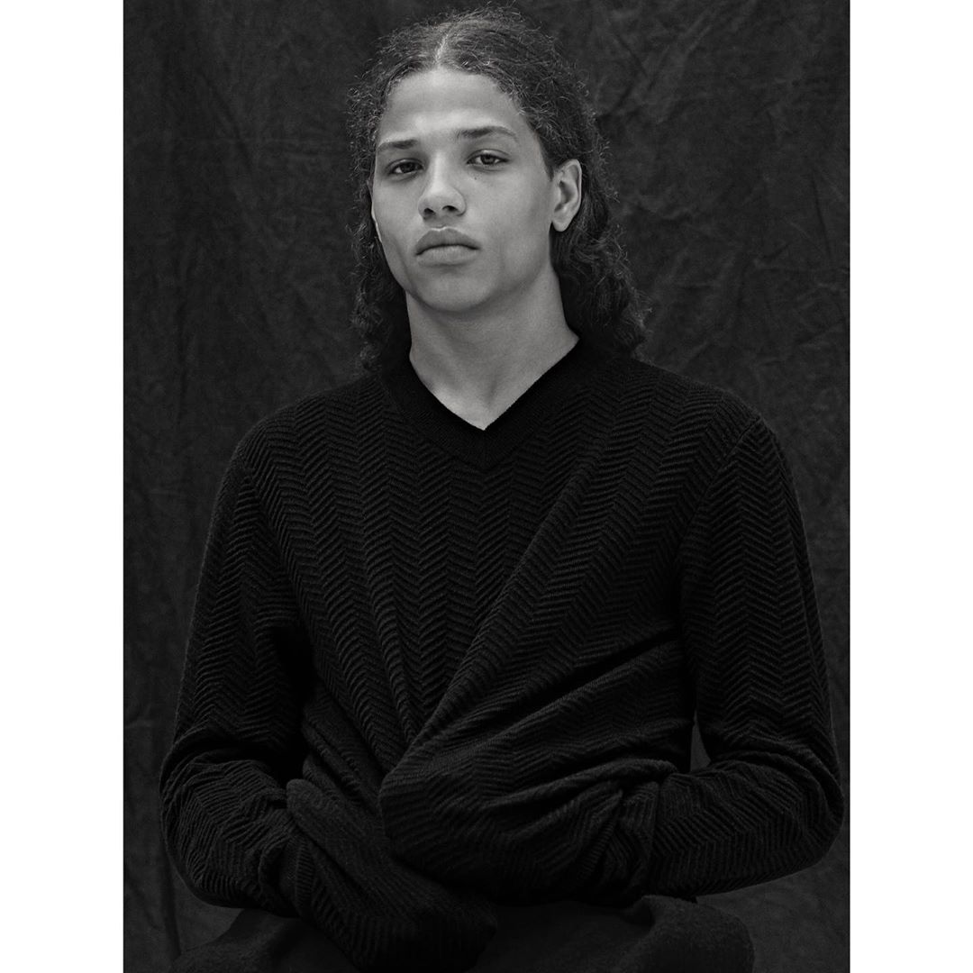 Monochrome portrait of a young adult with long curly hair, wearing a dark v-neck sweater, posing indoors against a textured backdrop.