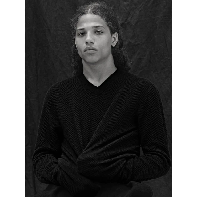 Monochrome portrait of a young adult with long curly hair, wearing a dark v-neck sweater, posing indoors against a textured backdrop.