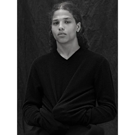 Monochrome portrait of a young adult with long curly hair, wearing a dark v-neck sweater, posing indoors against a textured backdrop.