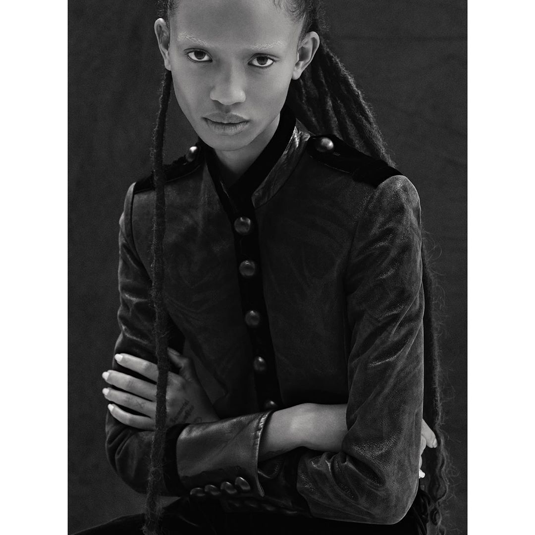 Portrait of a Black woman with dreadlocks, wearing a dark velvet jacket with button details, shot in black and white.