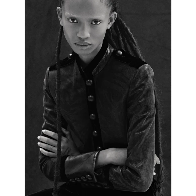 Portrait of a Black woman with dreadlocks, wearing a dark velvet jacket with button details, shot in black and white.