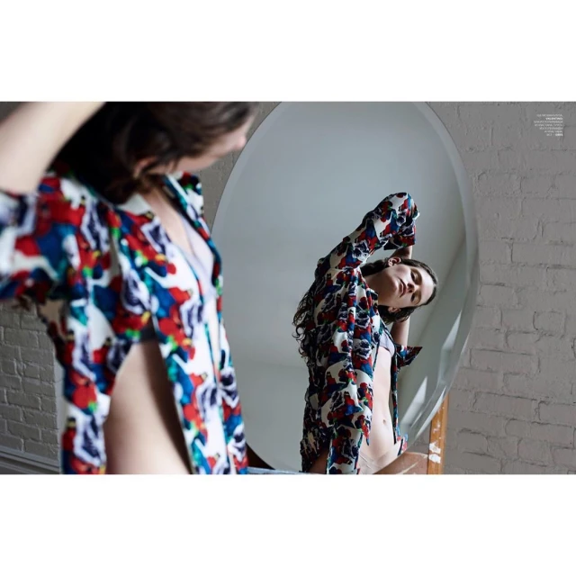 Indoor portrait of a woman with long brown hair wearing a patterned shirt, looking into a large oval mirror reflecting her image.