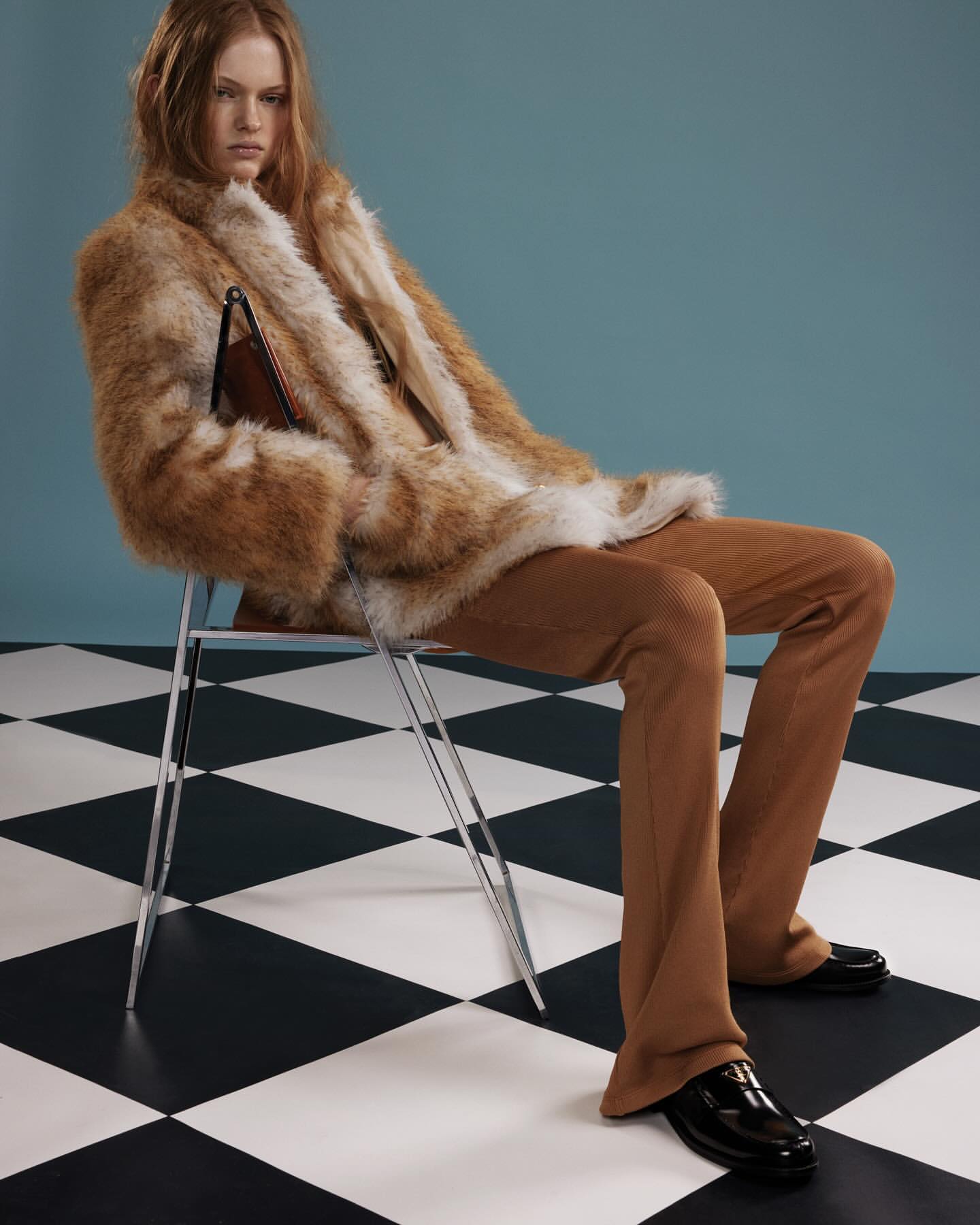 Fashion shot of a red-haired woman in a faux fur coat and corduroy pants sitting on a chair in a photo studio with a checkered floor.