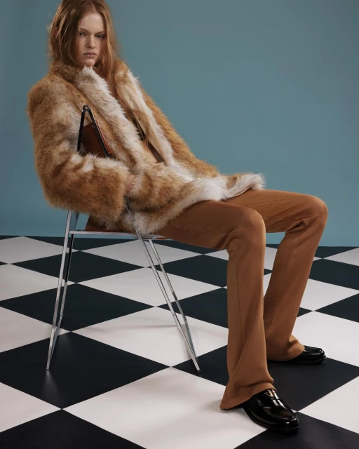 Fashion shot of a red-haired woman in a faux fur coat and corduroy pants sitting on a chair in a photo studio with a checkered floor.