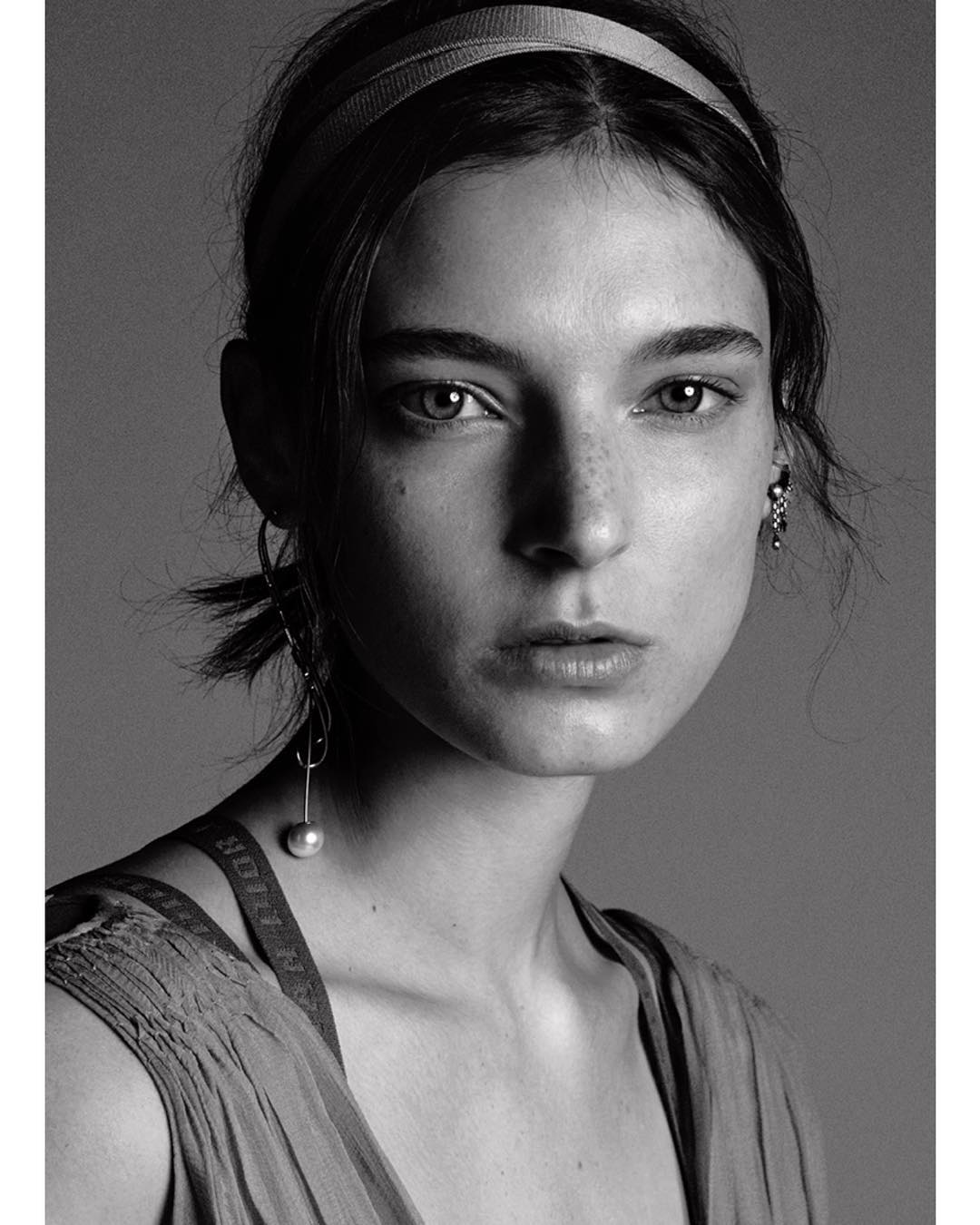 Monochrome portrait of a young woman with neutral expression, wearing a headband and drop earrings against a grey background.