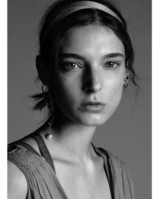 Monochrome portrait of a young woman with neutral expression, wearing a headband and drop earrings against a grey background.