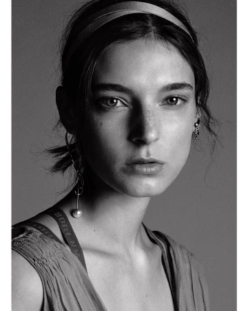 Monochrome portrait of a young woman with neutral expression, wearing a headband and drop earrings against a grey background.