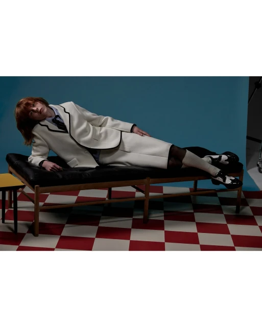 Woman in white suit with black trim lying on black bench in studio. Red and white checkered floor with blue backdrop.