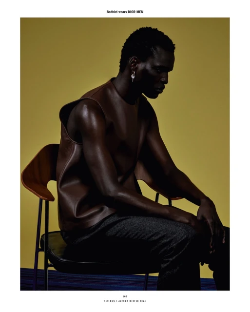 Dark-skinned male model with brown top sits on a chair against yellow backdrop for a fashion editorial. Studio lighting. 