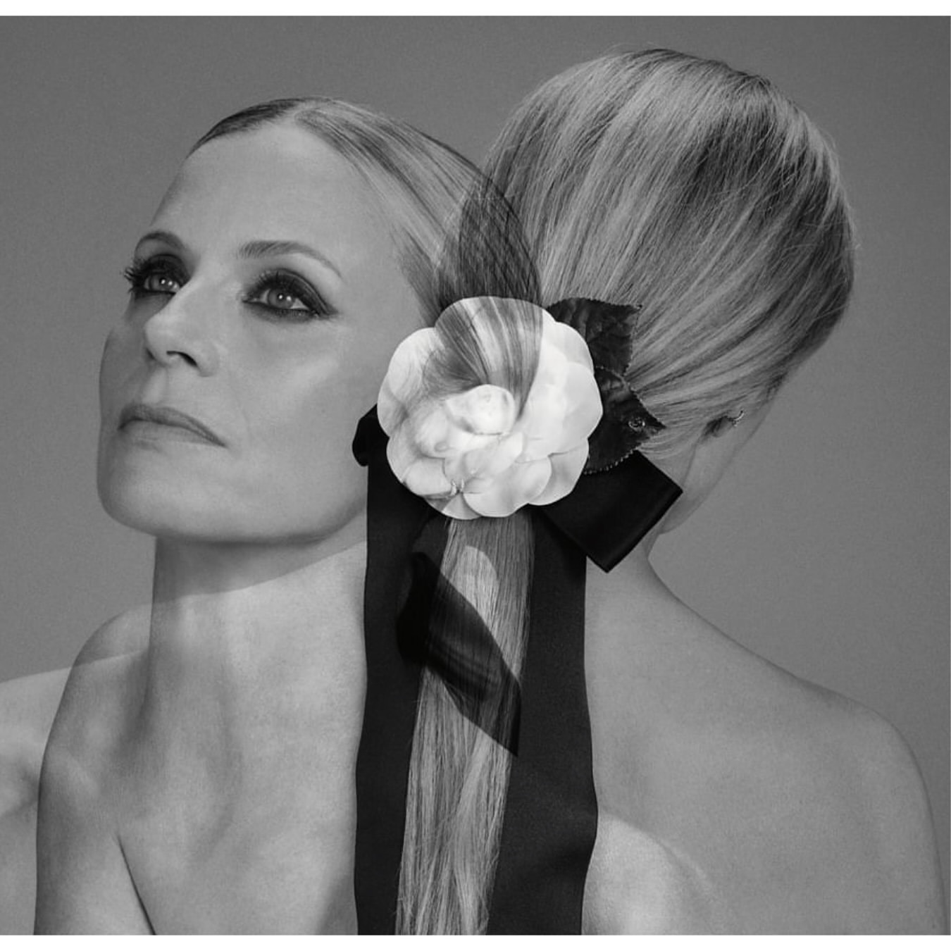 Black and white portrait of a woman with fair skin wearing a white flower hair accessory with a black ribbon and a neutral expression.