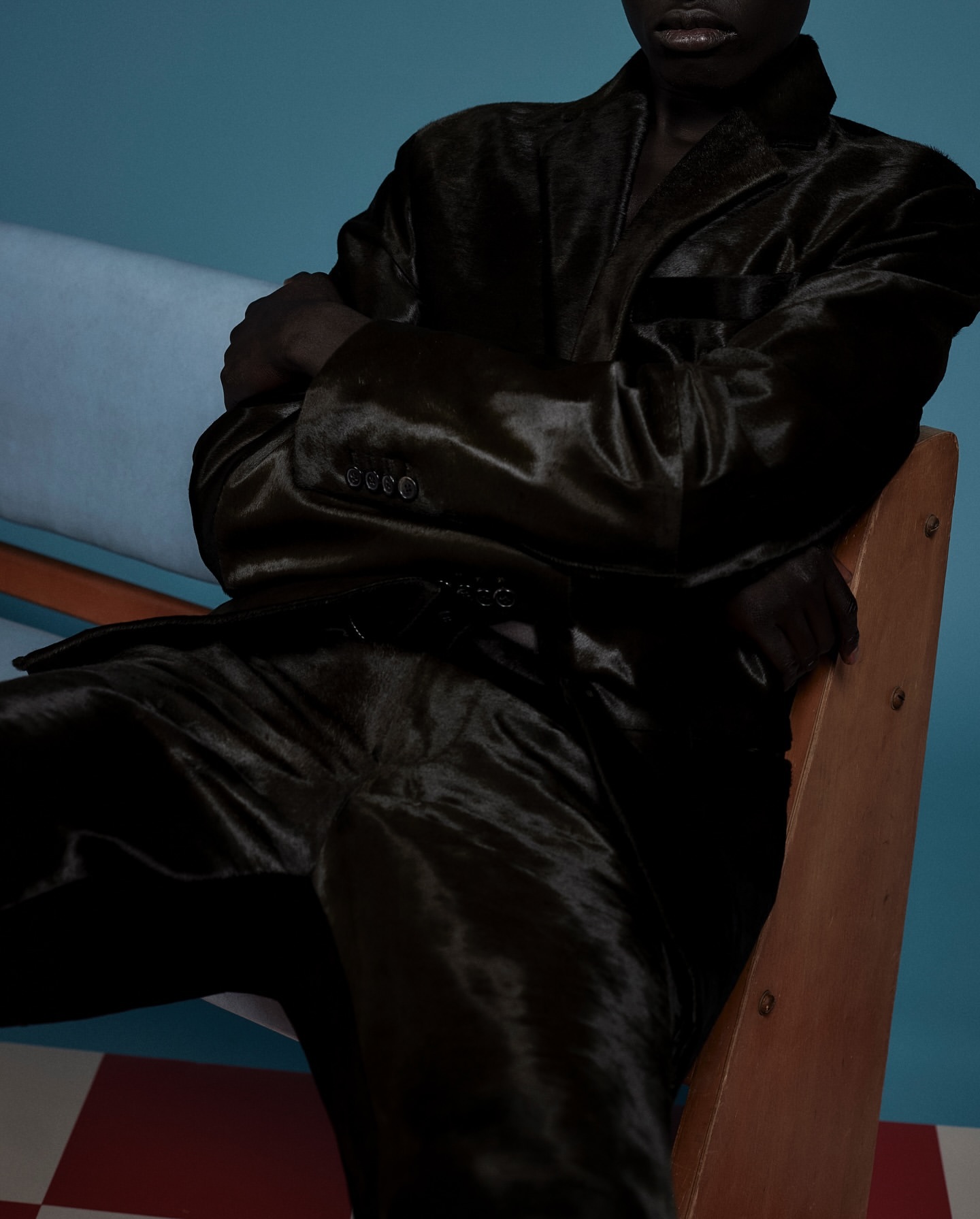 An adult with dark skin sits on a wooden chair wearing a dark shiny suit against a studio backdrop with a checkered floor.