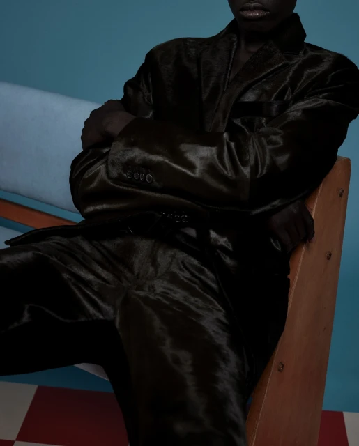 An adult with dark skin sits on a wooden chair wearing a dark shiny suit against a studio backdrop with a checkered floor.