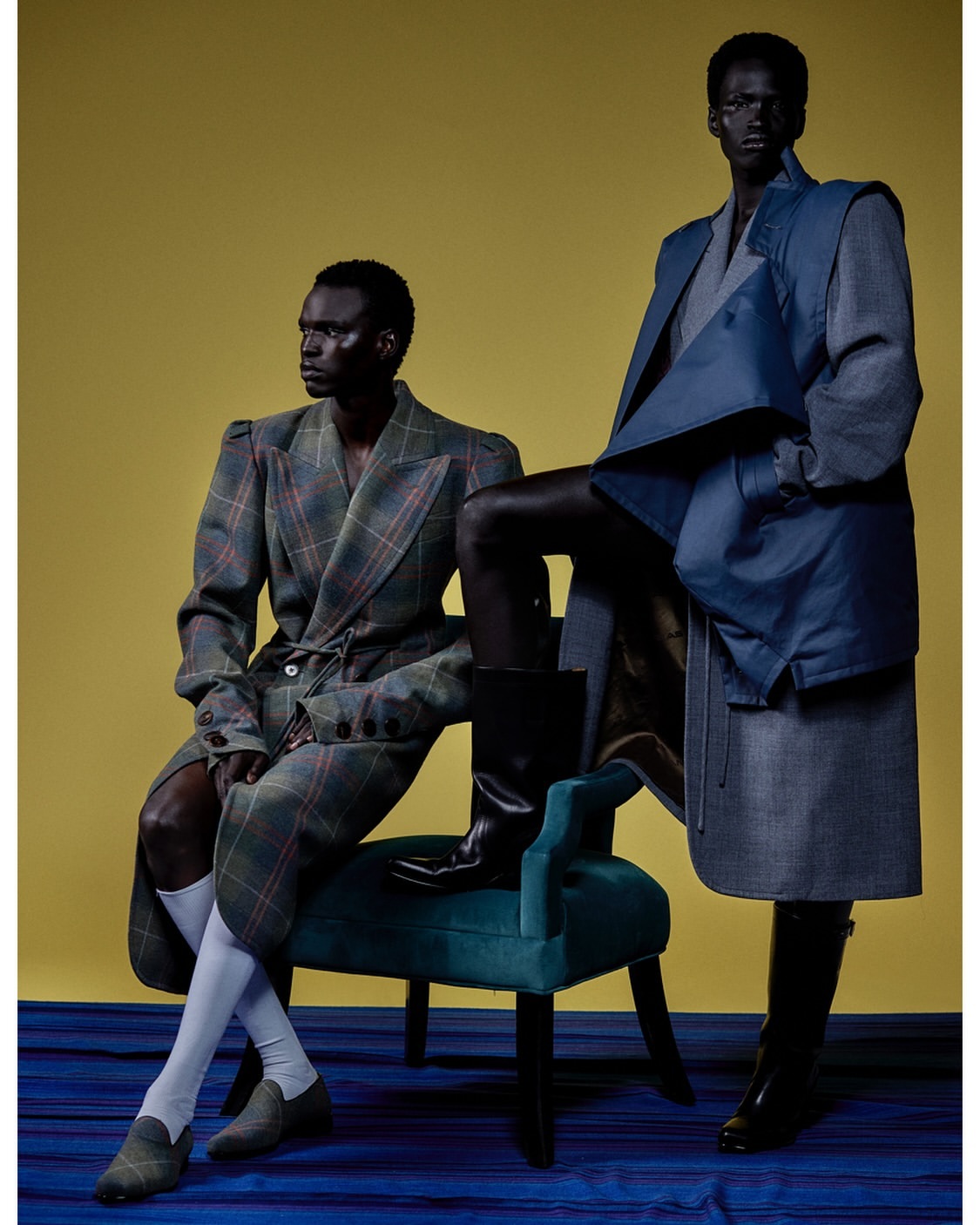 Two dark-skinned models in high-fashion attire pose against a yellow backdrop with a blue patterned floor, seated on a green velvet chair.