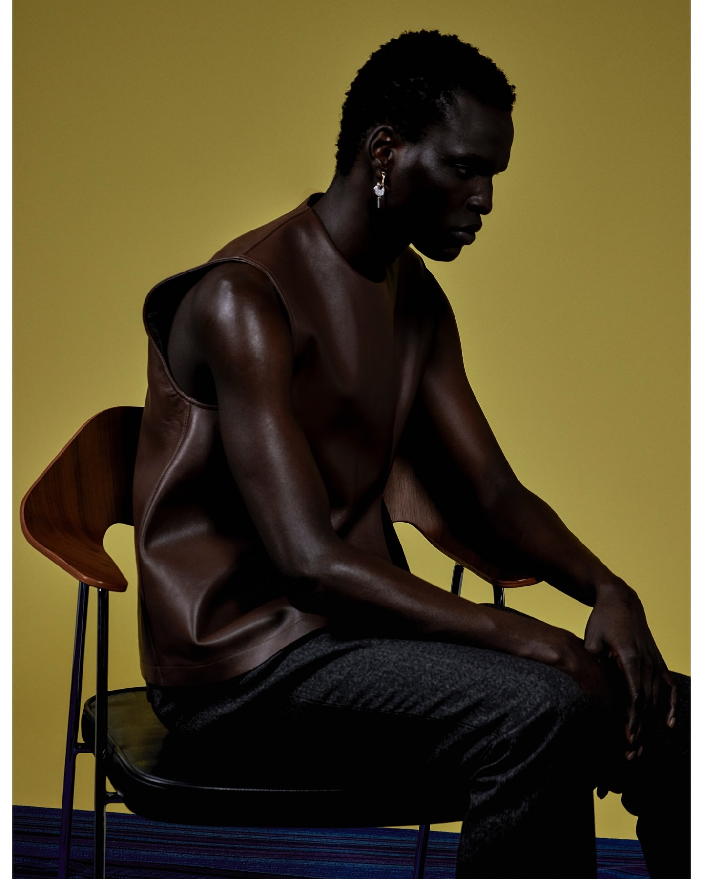 Dark-skinned adult male wearing brown leather sleeveless shirt sitting on a black chair against a yellow backdrop in a studio setting.