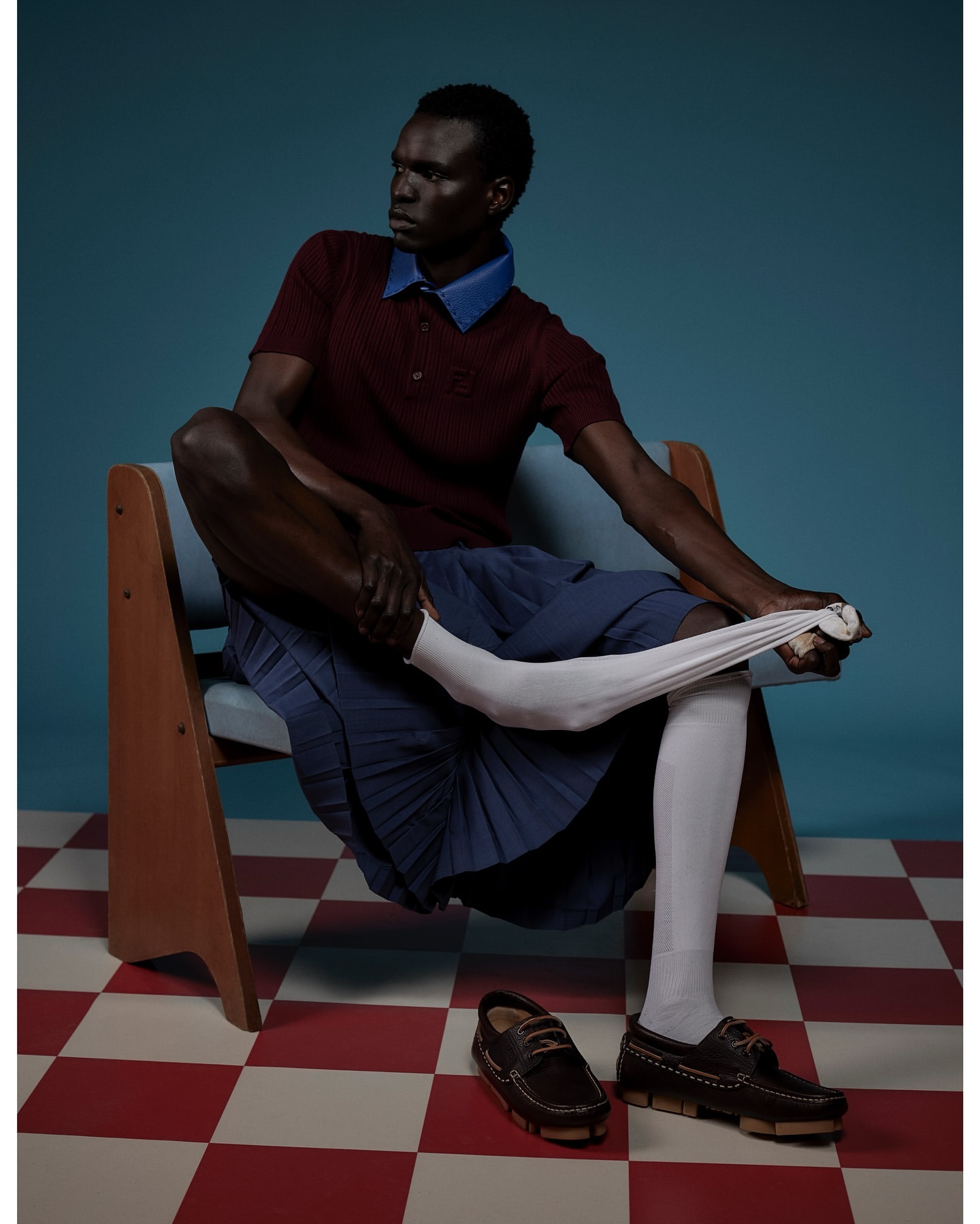 Editorial fashion shot of a Black male model seated and putting on a white sock, featuring a maroon polo shirt and navy skirt.