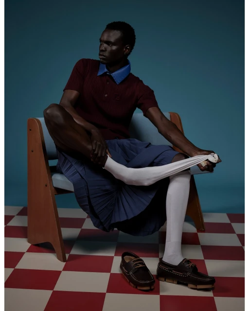 Editorial fashion shot of a Black male model seated and putting on a white sock, featuring a maroon polo shirt and navy skirt.