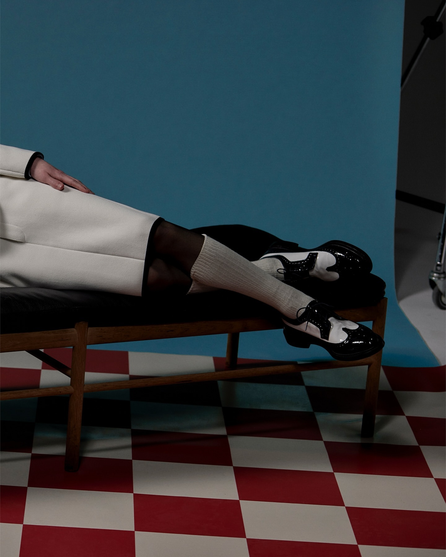 Person in white coat and sheer black tights sits on a wooden bench with black and white saddle shoes, posed against a blue backdrop.