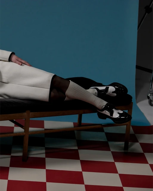 Person in white coat and sheer black tights sits on a wooden bench with black and white saddle shoes, posed against a blue backdrop.