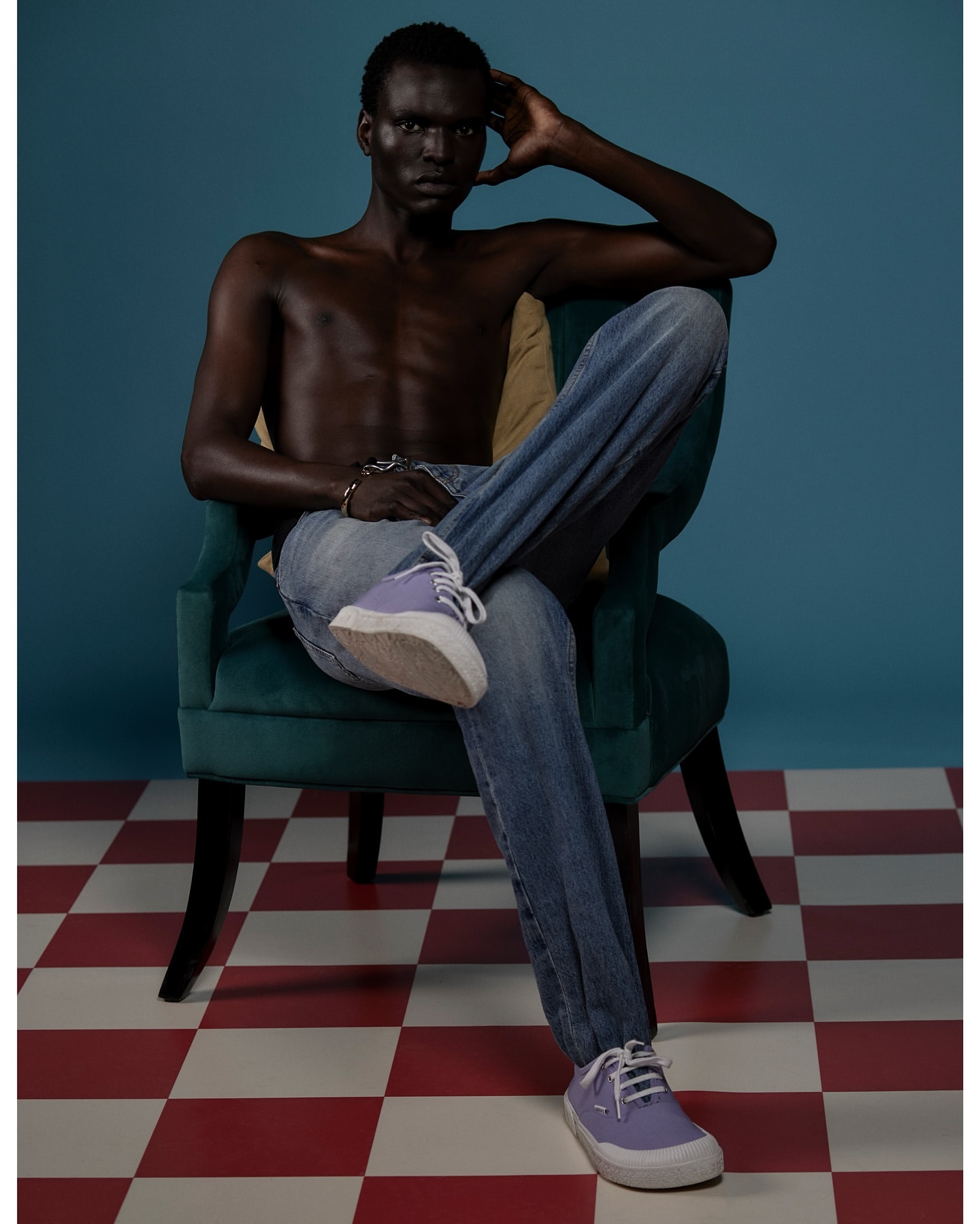 Shirtless man in blue jeans sits in a green velvet chair against a blue background, wearing purple sneakers on a checkered floor.