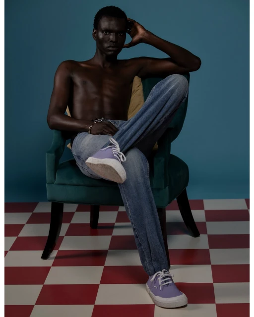 Shirtless man in blue jeans sits in a green velvet chair against a blue background, wearing purple sneakers on a checkered floor.