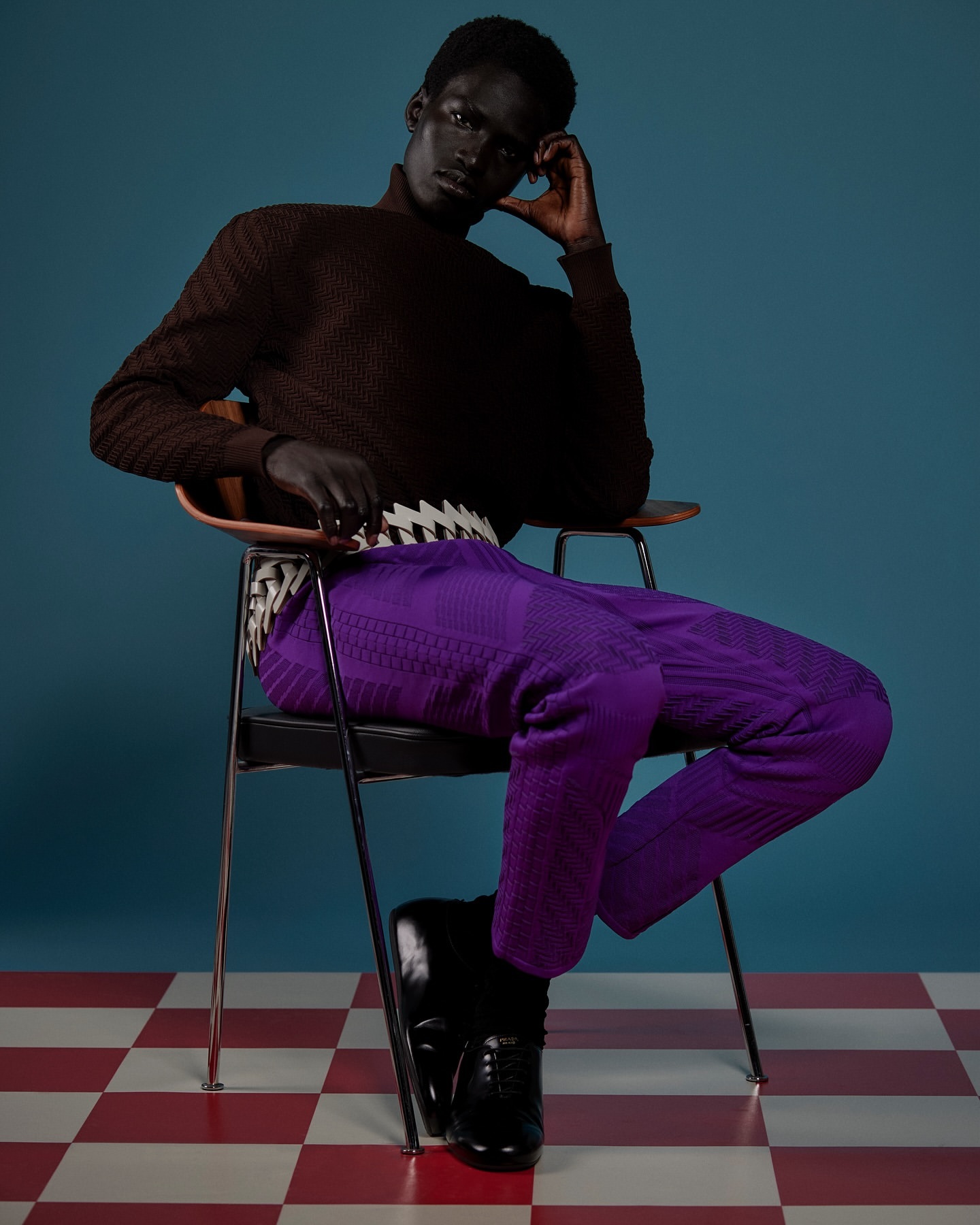 Stylish adult male model in a brown turtleneck and purple pants sits on a chair against a blue backdrop and red and white checkered floor.