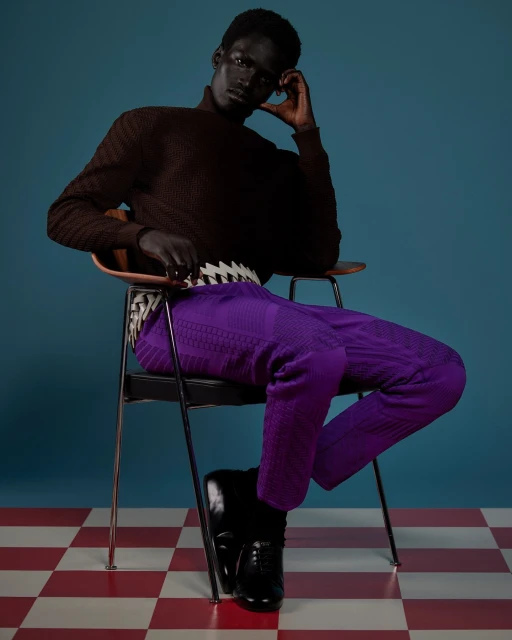 Stylish adult male model in a brown turtleneck and purple pants sits on a chair against a blue backdrop and red and white checkered floor.